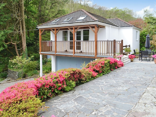 A white building with a wooden balcony, surrounded by pink flowers and stone paving.