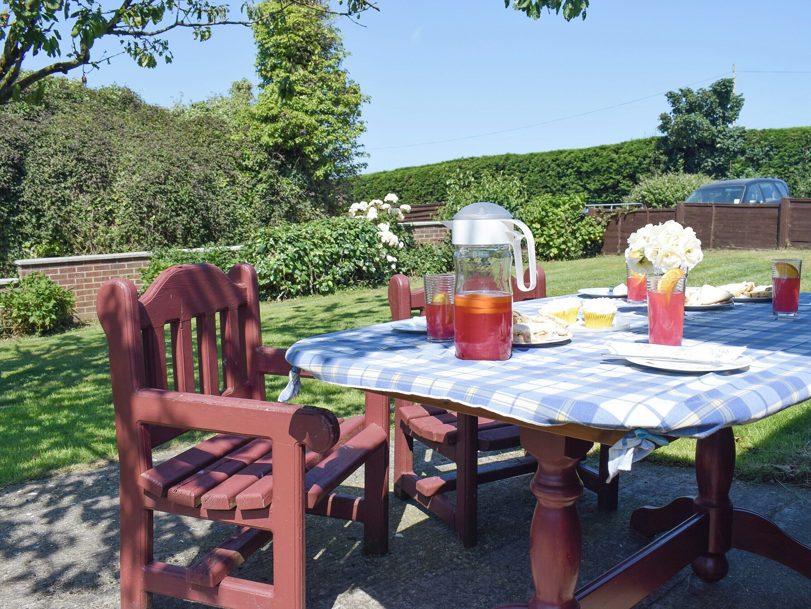 Une table de pique-nique recouverte d’une nappe à carreaux, entourée de chaises rouges. On y trouve des boissons, une carafe et une assiette remplie de nourriture.
