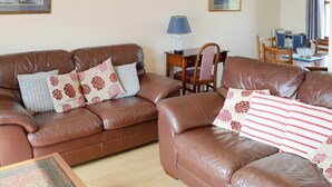 A living room with two brown leather sofas, a wooden side table, a lamp, and a framed picture on the wall. - Lochinchard Cottages (Kinlochbervie, Sutherland)