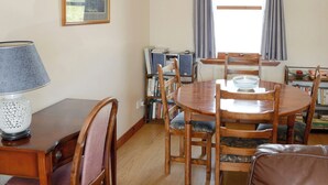 A dining area with a wooden table and chairs, a lamp, and a bookshelf with books. - Lochinchard Cottages (Kinlochbervie, Sutherland)