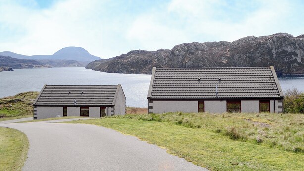 Two small, modern buildings with tiled roofs, situated on a grassy area beside a body of water, with mountains in the background. - Lochinchard Cottages (Kinlochbervie, Sutherland)