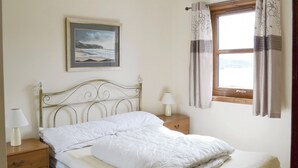 A neatly made bed with a headboard, a bedside table with a lamp, a wooden nightstand, and a window with curtains. - Lochinchard Cottages (Kinlochbervie, Sutherland)
