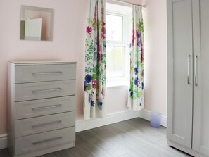 A room with a white wardrobe, a grey dresser, a window with floral curtains, and a small trash bin. - 2 bedroom accommodation in Nantlle, near Beddgelert (Nantlle, near Beddgelert)