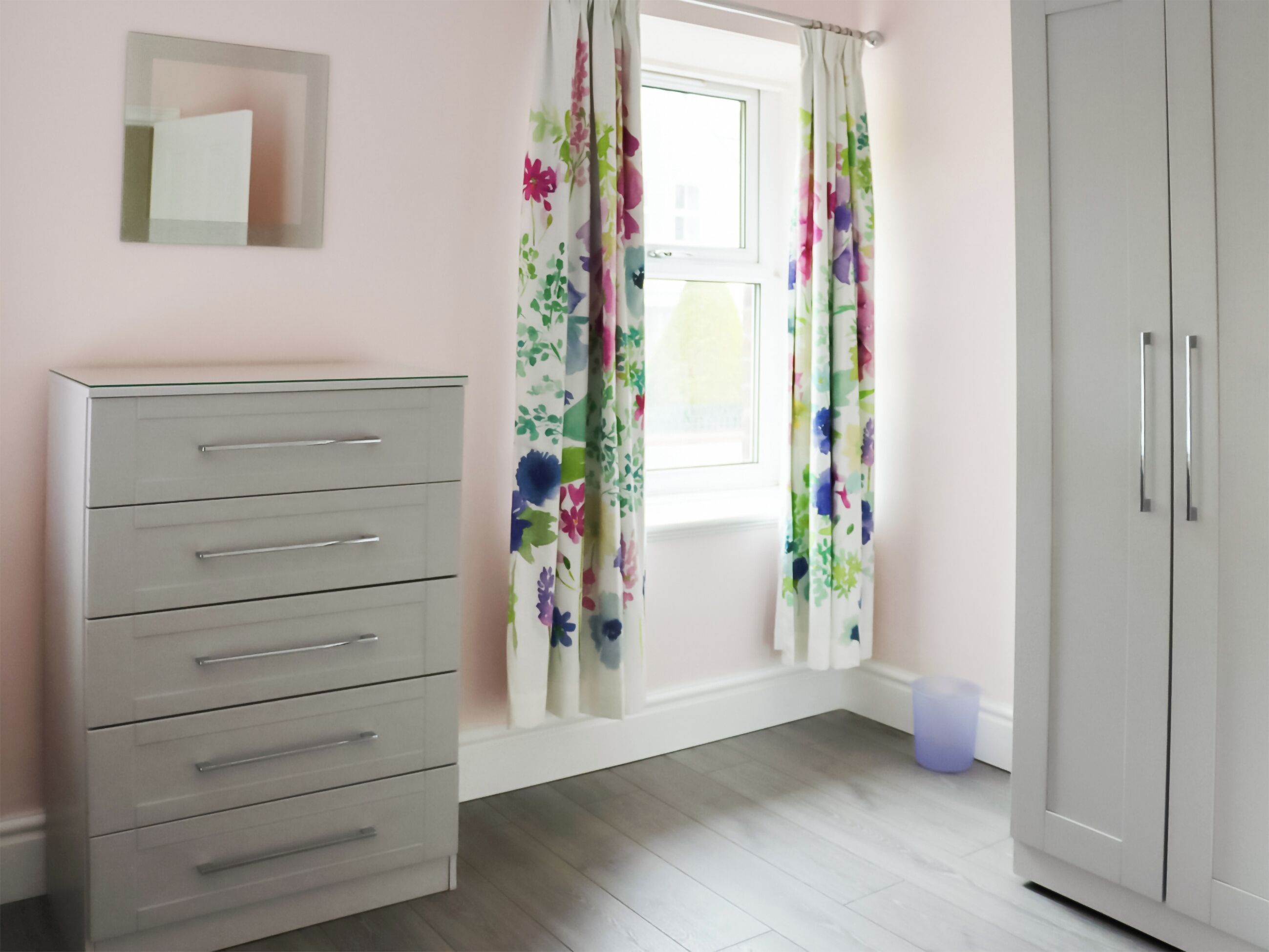 A room with a white wardrobe, a grey dresser, a window with floral curtains, and a small trash bin.