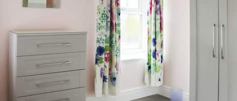 A room with a white wardrobe, a grey dresser, a window with floral curtains, and a small trash bin.