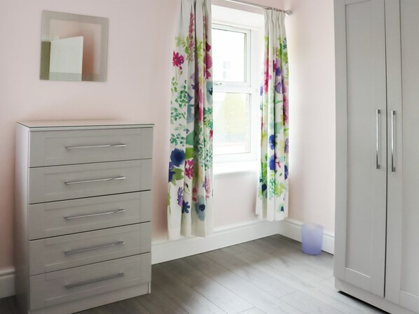 A room with a white wardrobe, a grey dresser, a window with floral curtains, and a small trash bin.