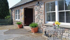 A stone building with a dark wooden door, large windows with white shutters, and potted plants in front.