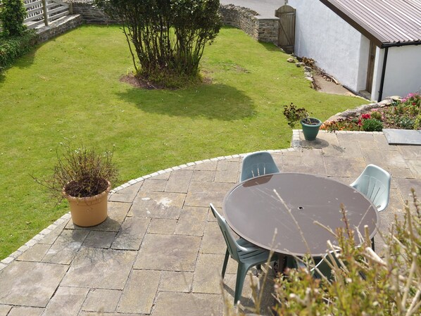 Een terras met een stenen tafel en twee stoelen, omgeven door een verzorgd gazon en een stenen muur.