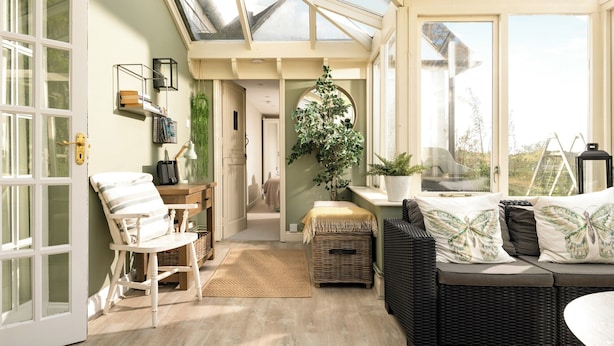 Habitación soleada con techo alto, sofá de mimbre, escritorio de madera y vista al exterior a través de amplias ventanas.
