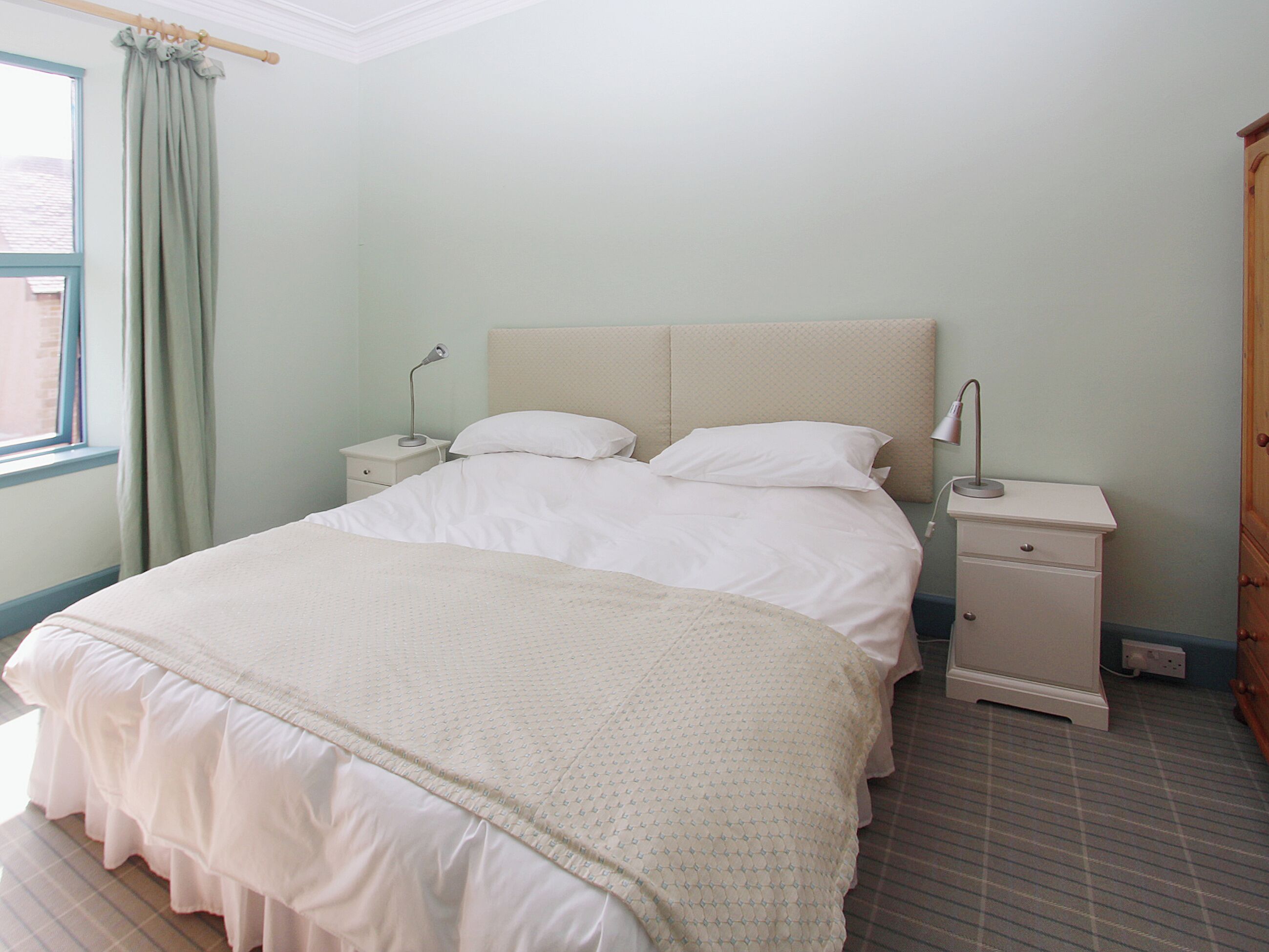 Un dormitorio con una cama, dos mesitas de noche, una ventana con cortinas y una puerta.