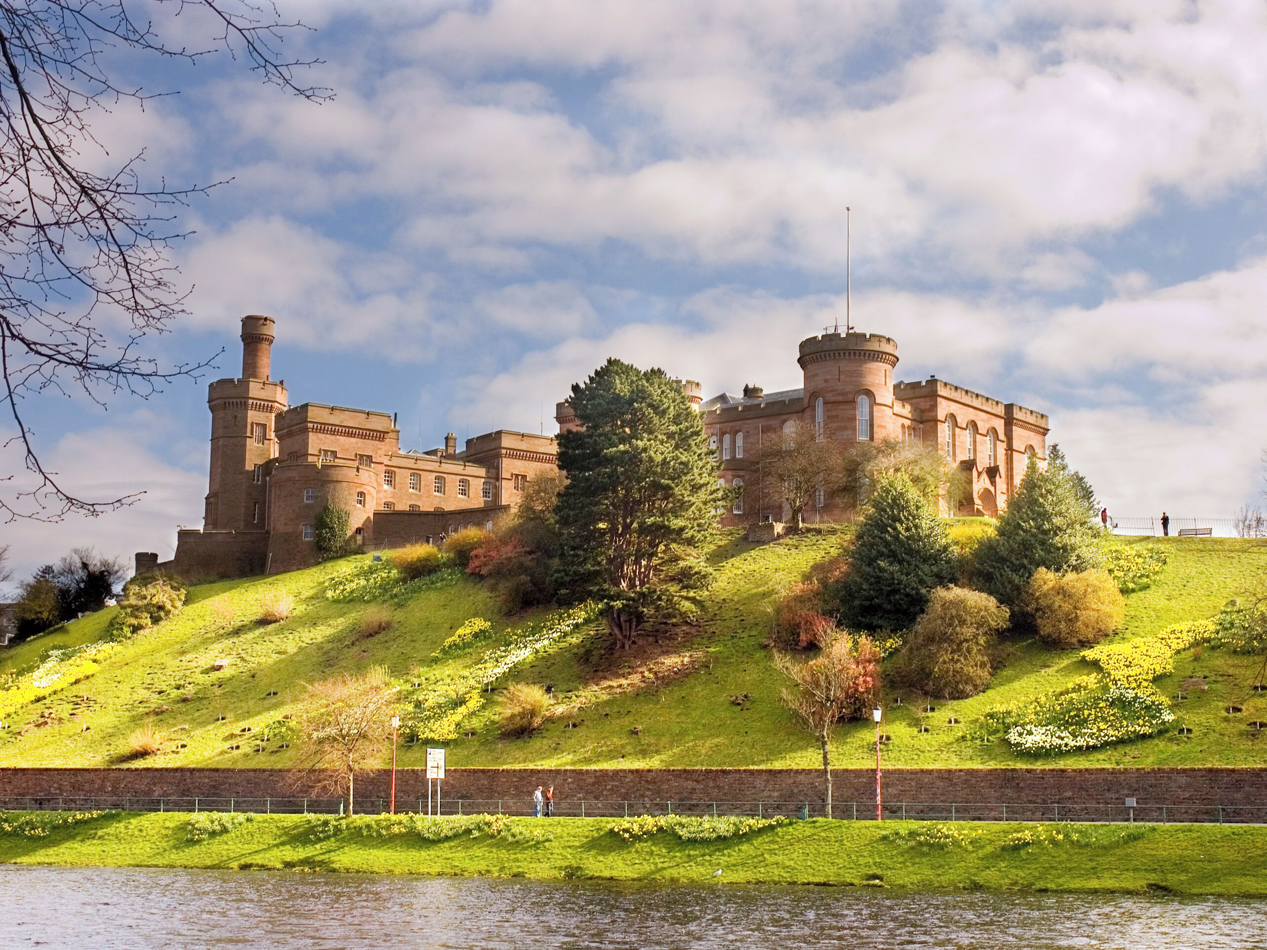 A large castle-like building with multiple towers, situated on a grassy hill with a body of water in the foreground.