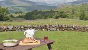 A picnic setup with a table, food, and drink, overlooking a scenic landscape with hills and sheep. - Birkerthwaite Farmhouse (Eskdale)
