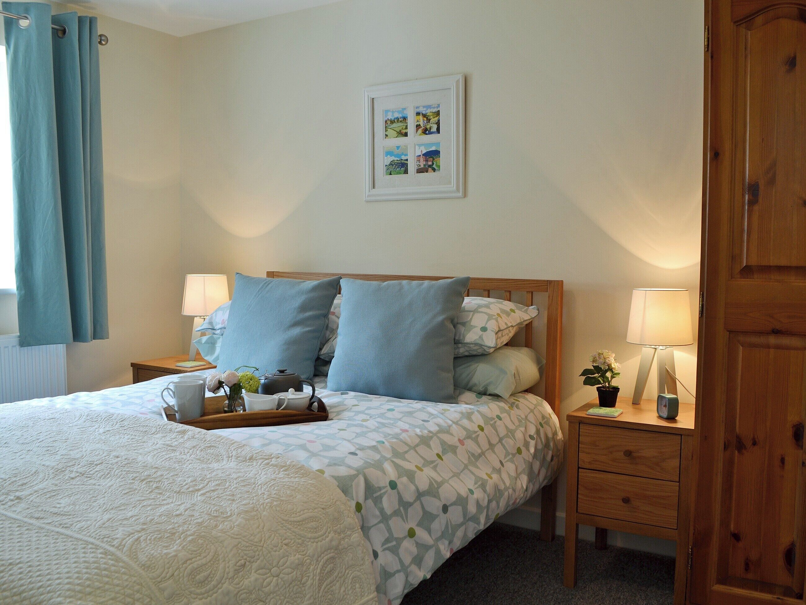 A neatly arranged bedroom with a bed, two bedside tables, a nightstand, a wooden wardrobe, and a wall-mounted picture.