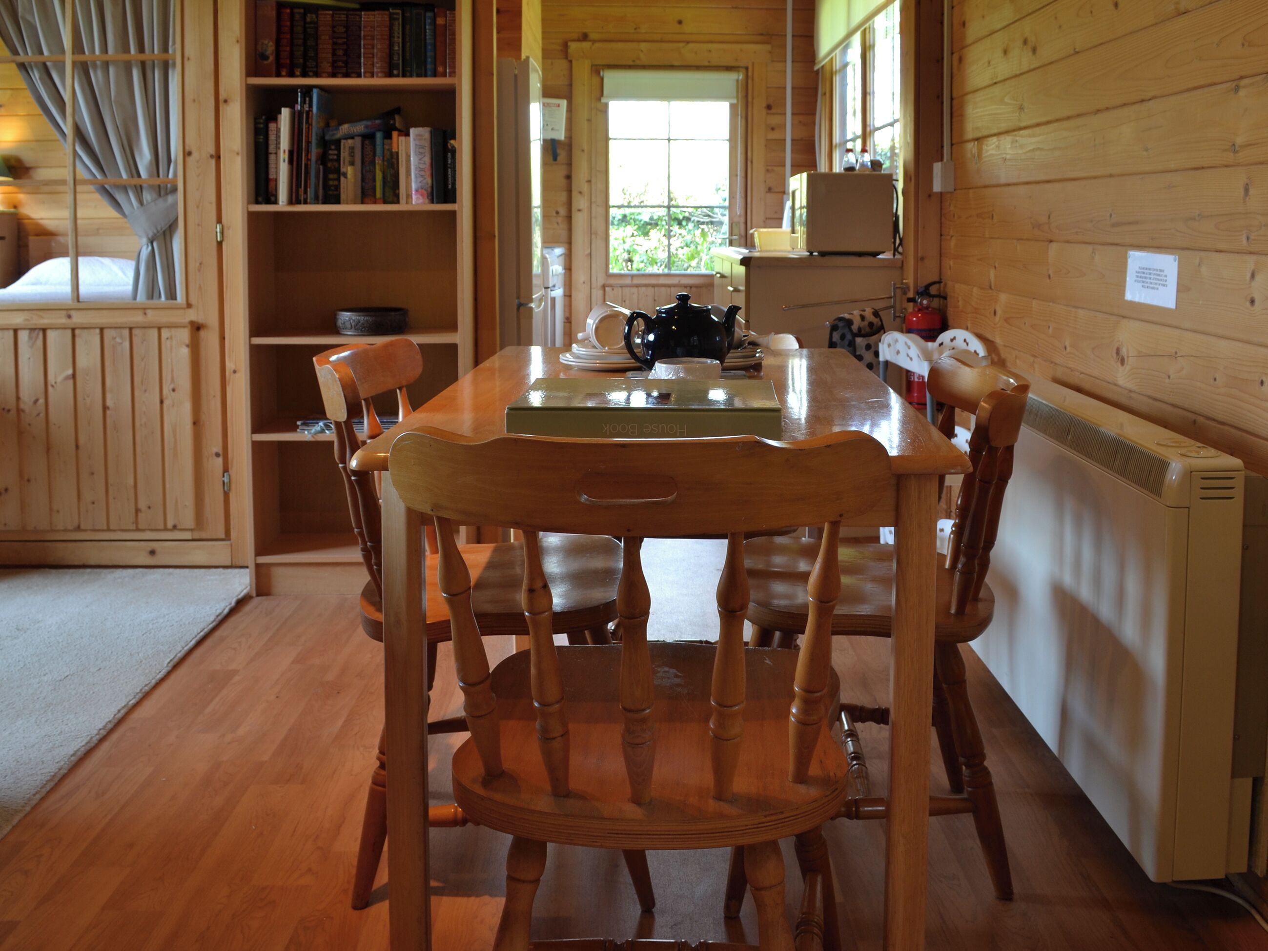A wooden dining table with chairs, a bookshelf, and a window with a view of greenery.