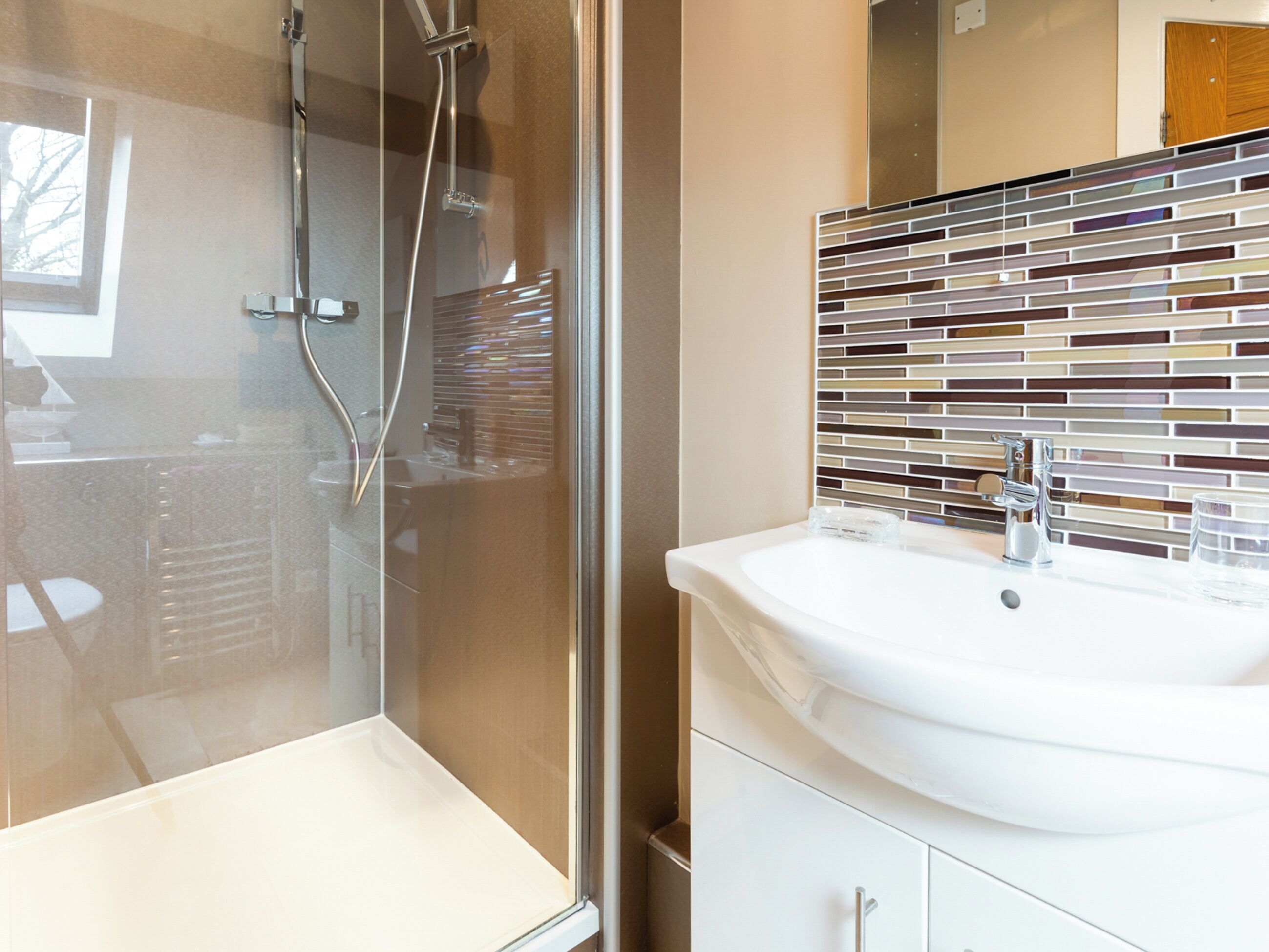 Un baño con ducha envuelta en vidrio, un lavamanos blanco con grifo moderno y un salpicadero de azulejos.