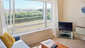 A room with a large window, a sofa, a TV on a stand, and a remote control on a small table. - Brightlands Apartment (Bude)