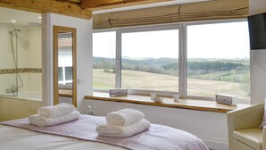A bedroom with a large bed, a window with a view of the countryside, a shower, and a bedside table with towels.