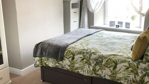 Een slaapkamer met een bed, nachtkastje, dressoir en een raam met gordijnen.