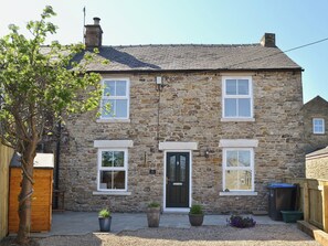 A stone house with a black door, two windows on the ground floor, and a small porch area with potted plants.