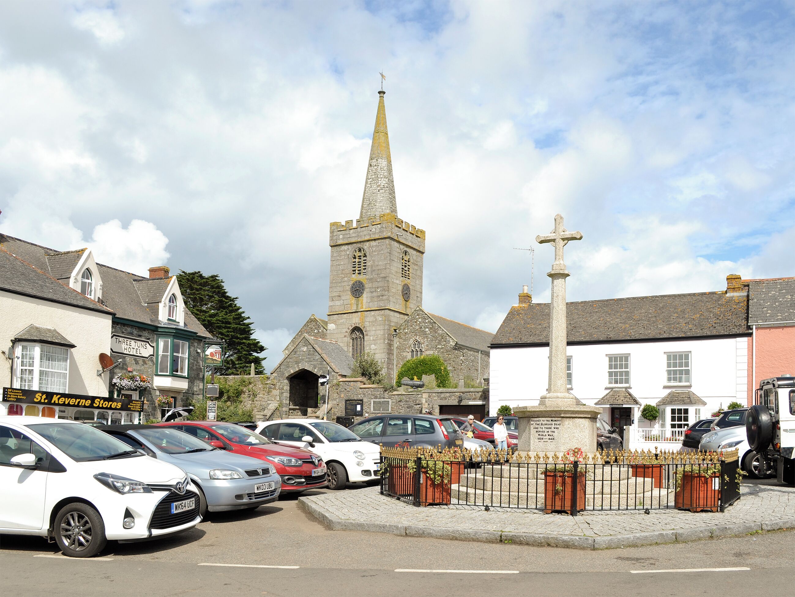 A church with a tall spire, a white building with a cross, and a parking area with cars.