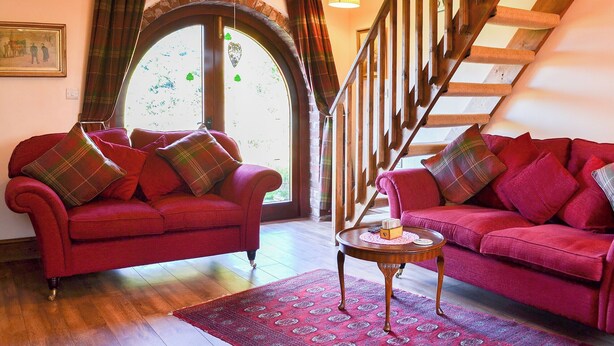A cozy living room with two red sofas, a wooden staircase, and a large arched window. - 3 bedroom accommodation in Laytham, near York (Laytham, near York)