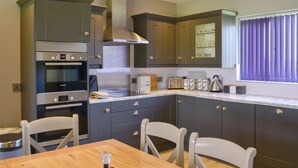 A modern kitchen with built-in appliances, a dining area with a wooden table and chairs, and a window with blinds. - Lucy's Lodge - UKC2202 (Doddington, near Lincoln)