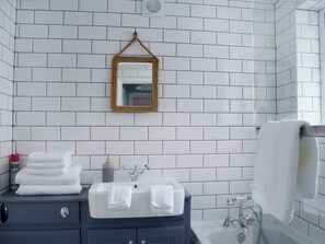 Et badeværelse med hvide flisevægge, en blå kommode med vask, to håndklæder, et spejl med gylden ramme og et bruseområde.