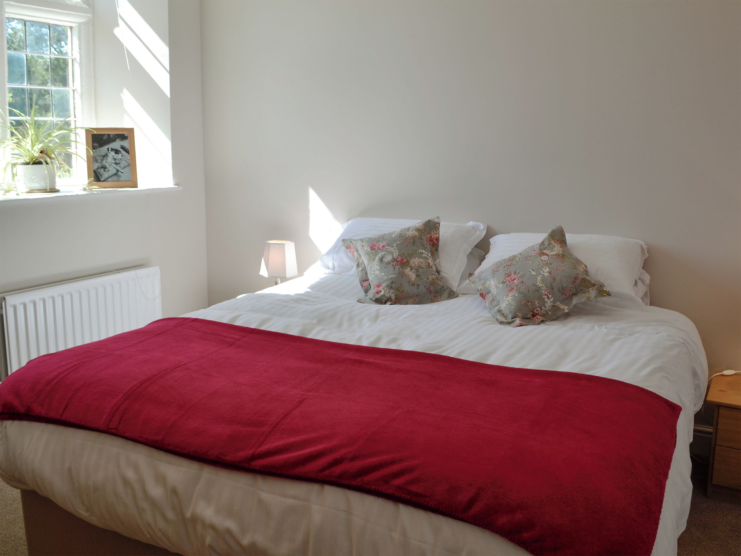 Un lit bien fait, recouvert d’une couverture rouge, avec deux coussins à motifs floraux et un tableau encadré posé sur la table de chevet.