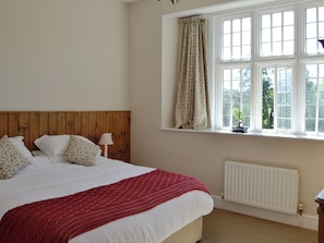 A bedroom with a wooden headboard, a bed with white bedding and a red blanket, a window with curtains, and a radiator.