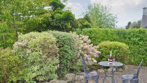 Ein Garten mit einer gepflasterten Terrasse, einem runden Tisch und vier Stühlen, umgeben von verschiedenen Sträuchern und Bäumen.