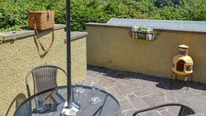A patio with a table, chairs, a wicker chair, a glass table, a metal umbrella, a wicker basket, a wall-mounted bird feeder, and a view of hills. - 2 bedroom accommodation in Llandygai, near Bangor (Llandygai, near Bangor)