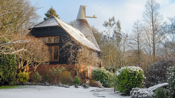 Una casa tradicional con techo inclinado, chimenea y jardín cubierto de nieve.