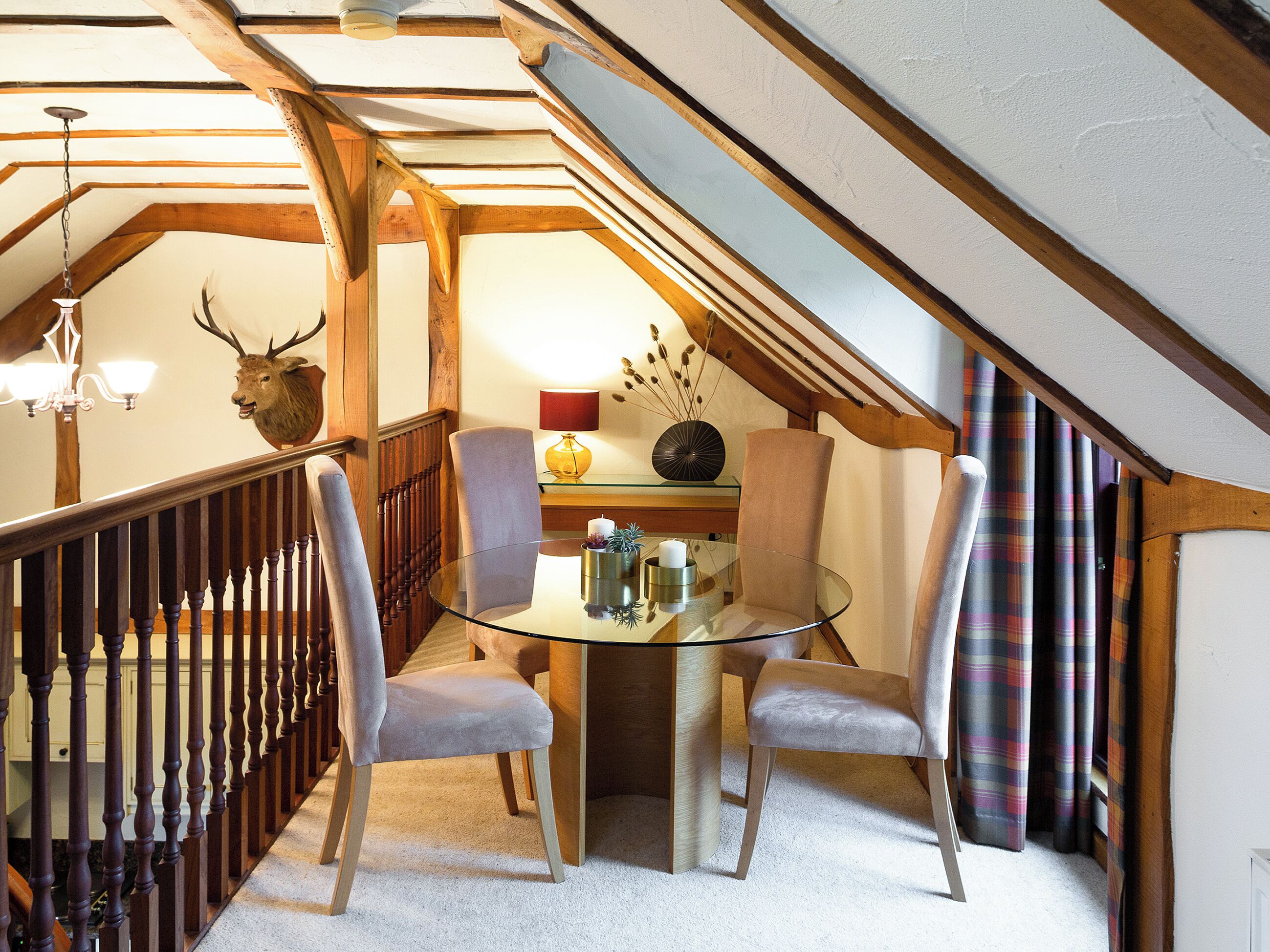 A dining area with a glass table, two chairs, a chandelier, and a deer head mounted on the wall.