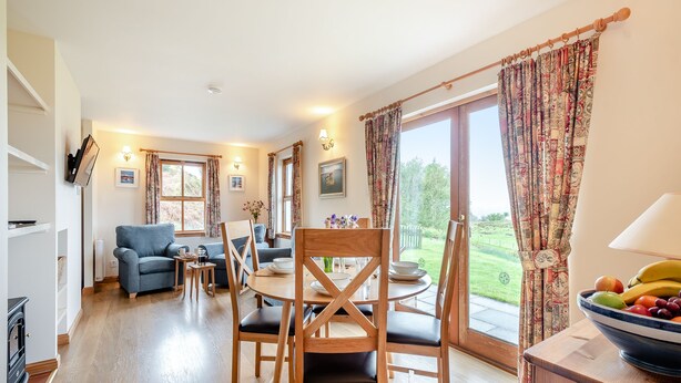 A cozy living room with a sofa, dining table, and chairs, a TV mounted on the wall, and a view of the outdoors through large windows. - 2 bedroom accommodation in Achnamara, near Lochgilphead (Achnamara, near Lochgilphead)