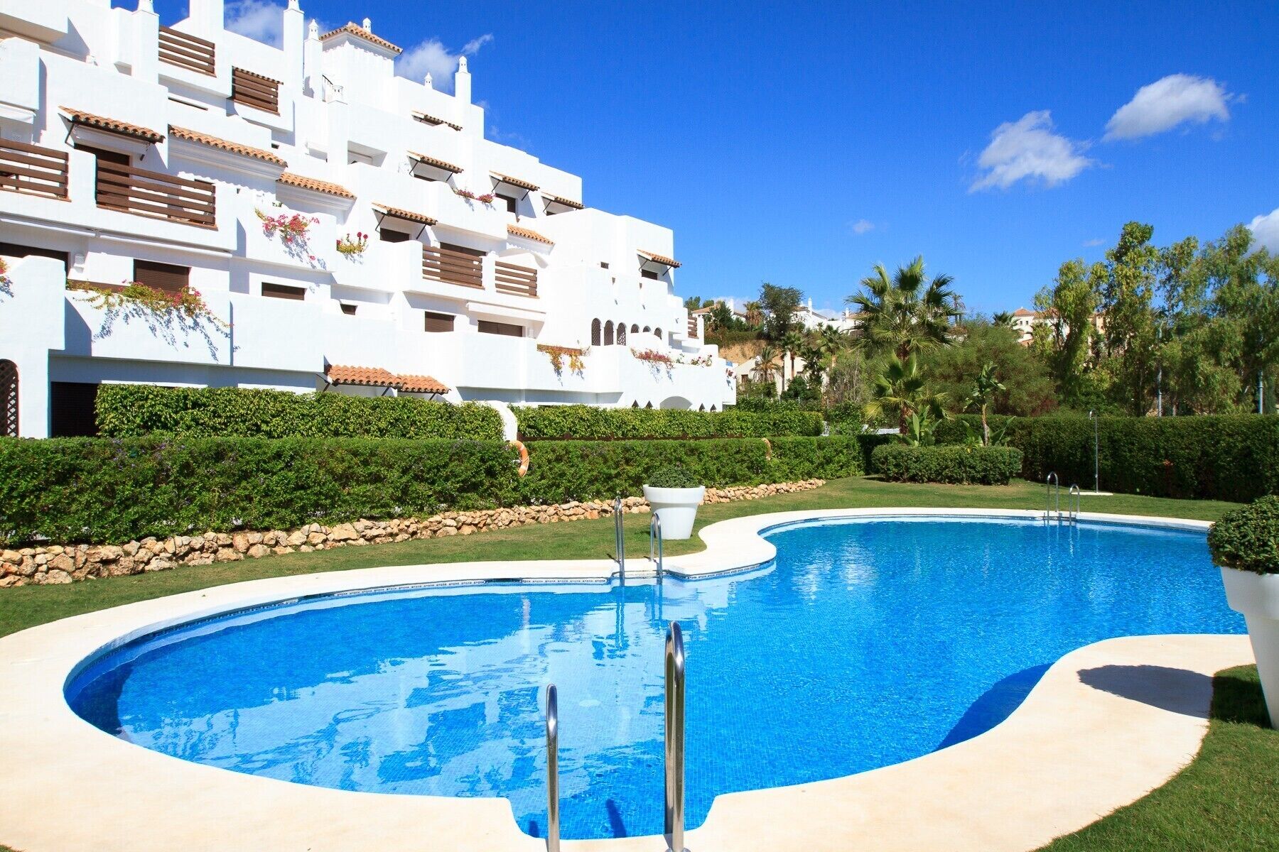 A swimming pool with a curved edge, surrounded by a well-maintained lawn and a stone wall. In the background, there is a white building with multiple balconies and a tiled roof.