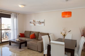 A modern living room with a sofa, coffee table, dining table, and chairs. There is a painting on the wall and a window with curtains.