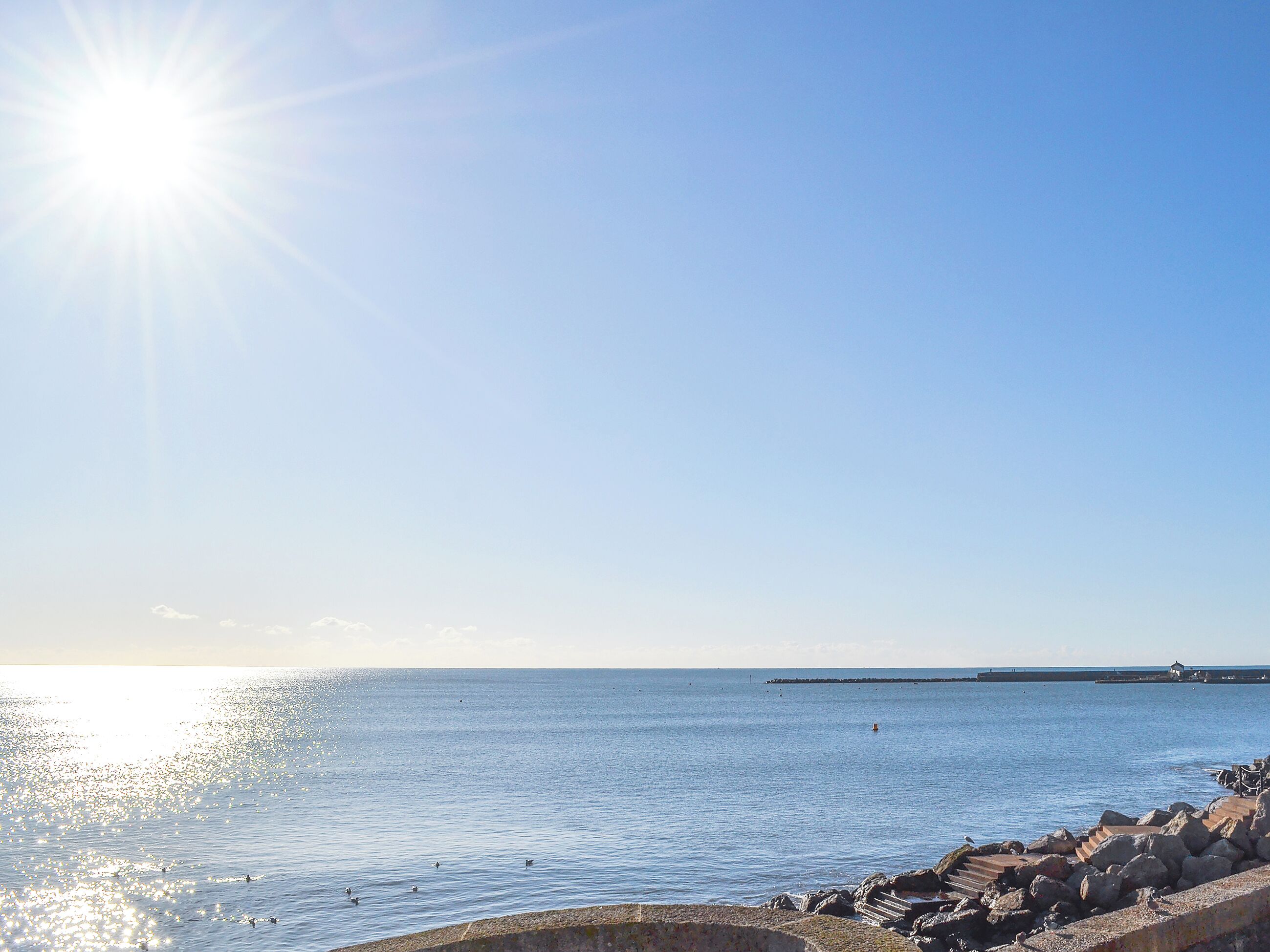 A coastal scene with a clear sky, calm sea, and a rocky breakwater.