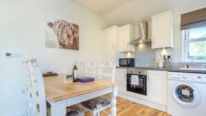 Een keuken met een houten tafel, stoelen, een magnetron, oven, wasmachine en een schilderij aan de muur.