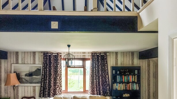 Een loft-achtig vertrek met een schuine kap, een houten trap, een groot raam met gordijnen, een bank, een lamp en een boekenkast.