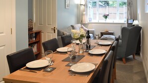 Un coin repas avec une table en bois dressée pour quatre personnes, entourée de chaises. On aperçoit un canapé et une fenêtre avec des rideaux au fond.
