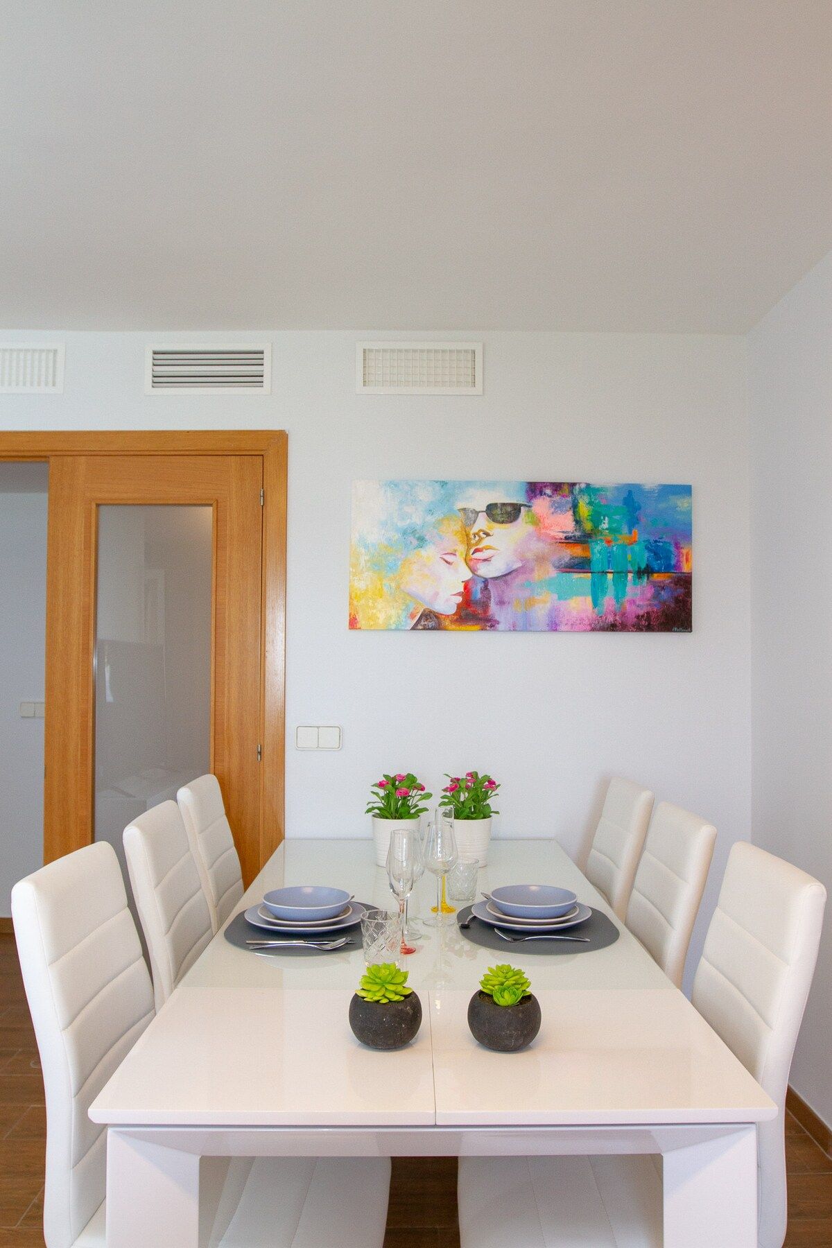 Un comedor con una mesa blanca, seis sillas, dos plantas en macetas y un cuadro abstracto colorido en la pared.