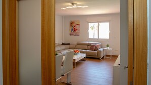 Ein Wohnzimmer mit einer Couch, einem Esstisch und einem kleinen weißen Tisch mit einer Pflanze. An der Wand hängt ein Gemälde und das Fenster bietet einen Blick ins Freie.