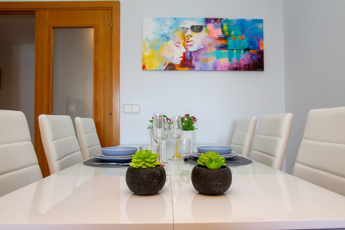 A dining table with four chairs, two potted plants, a colorful abstract painting, and a vase with flowers.