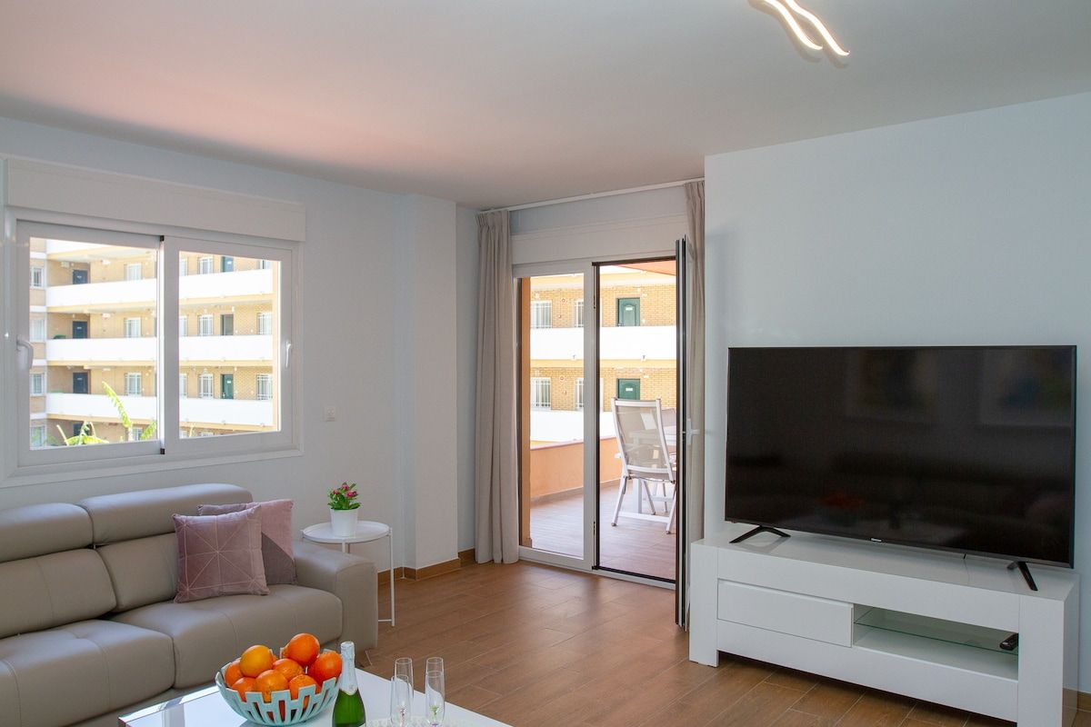 A modern living room with a sofa, a TV, and a balcony with a view of buildings.
