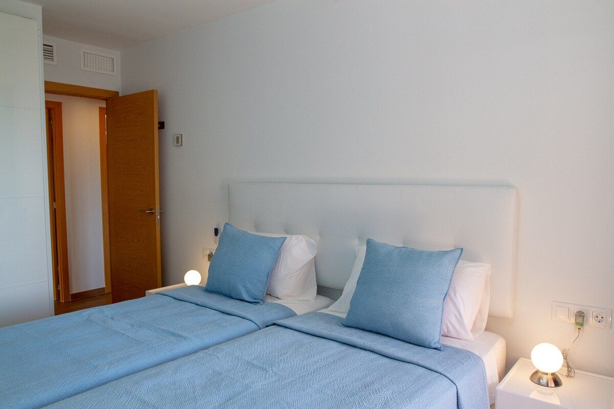 A double bed with a headboard, two bedside lamps, and a door leading to another room.