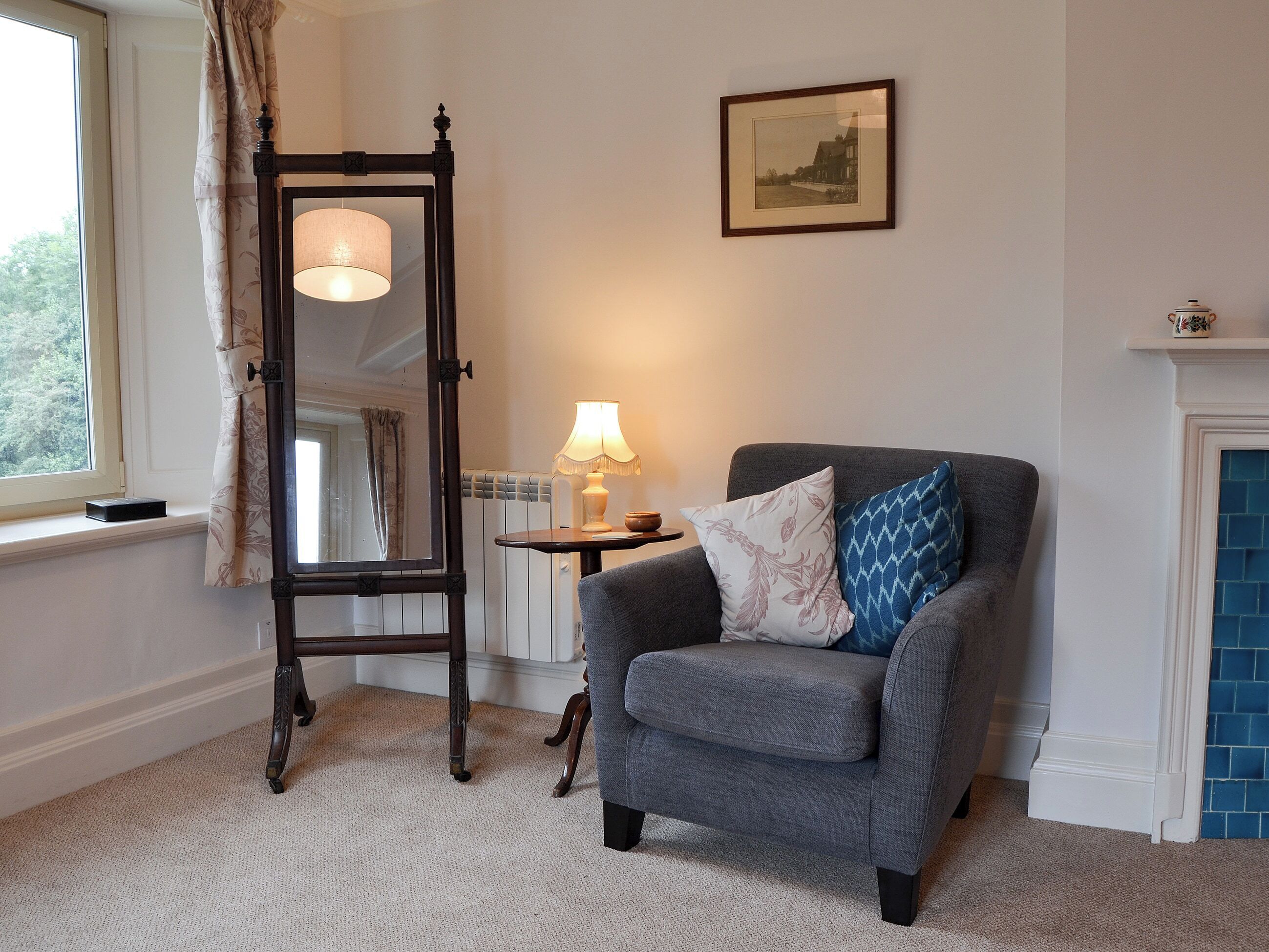 Een kamer met een grijze fauteuil, een staand spiegel, een tafel met een lamp en een lijst met een afbeelding aan de muur.