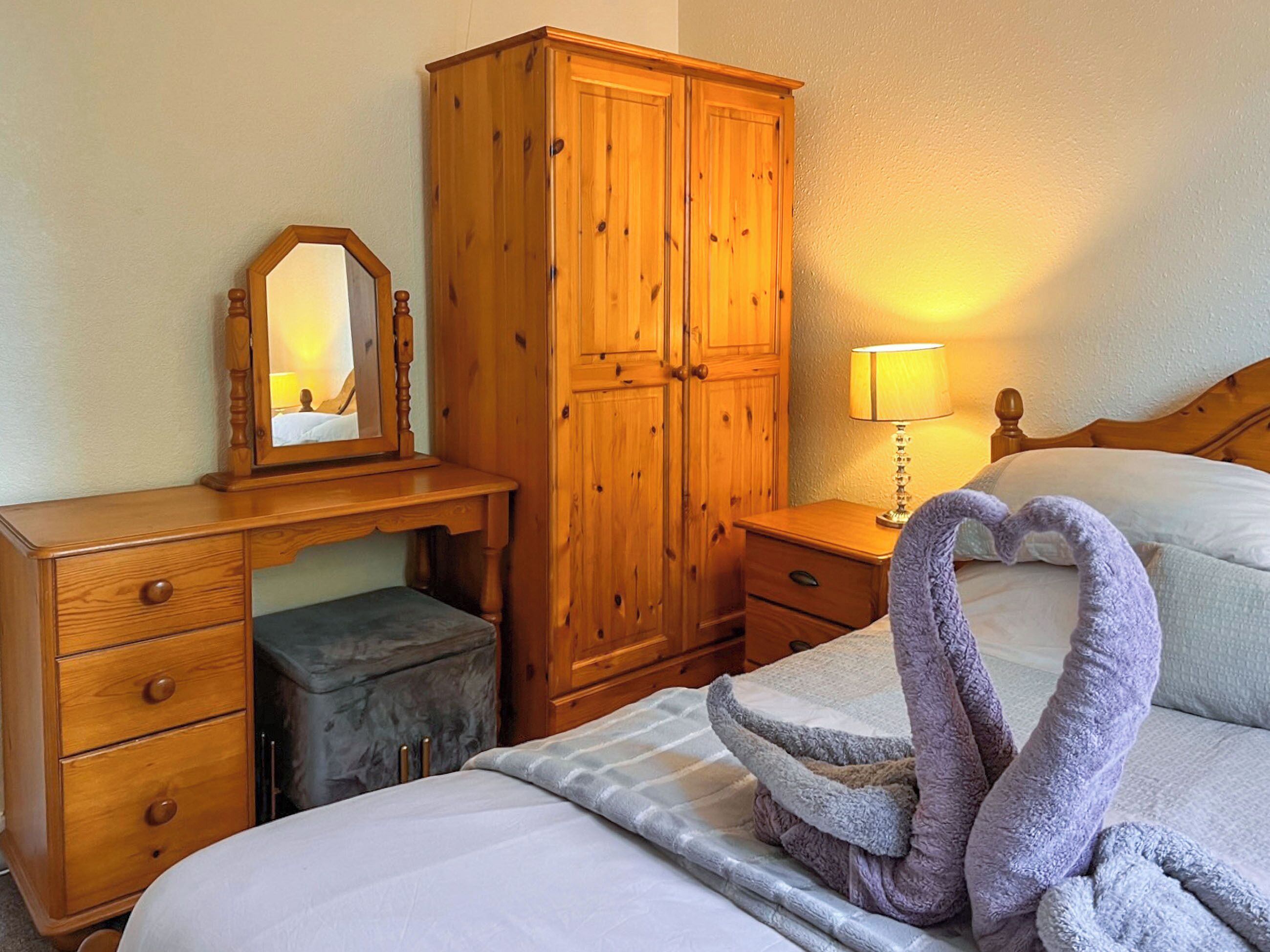 A hotel room with a wooden wardrobe, a desk with a mirror, a bedside table with a lamp, and a bed with a decorative towel arrangement.