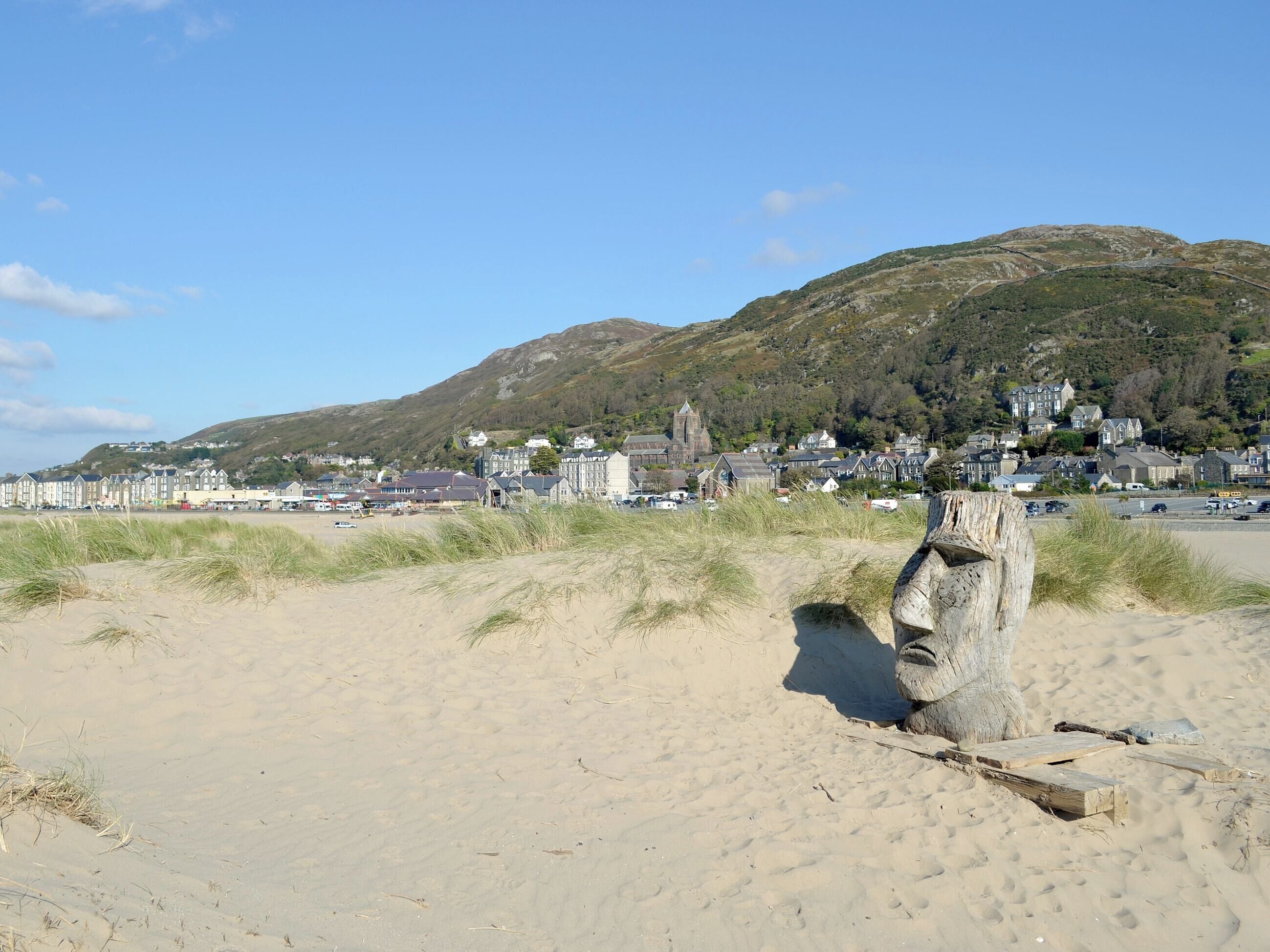 A sandy beach with a wooden structure, a town in the background, and a hill with buildings.