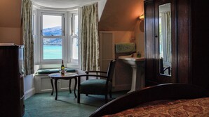 Ein Hotelzimmer mit Meerblick, ein Bett, ein Sessel, ein kleiner Tisch mit Flasche und Gläsern sowie ein Badezimmer mit einem Handtuch an der Wand.