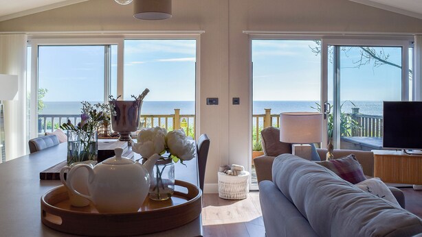 A spacious living area with a large window overlooking the sea, a dining table with a vase of flowers, and a comfortable sofa. - 2 bedroom accommodation in Corton, near Lowestoft (Corton, near Lowestoft)